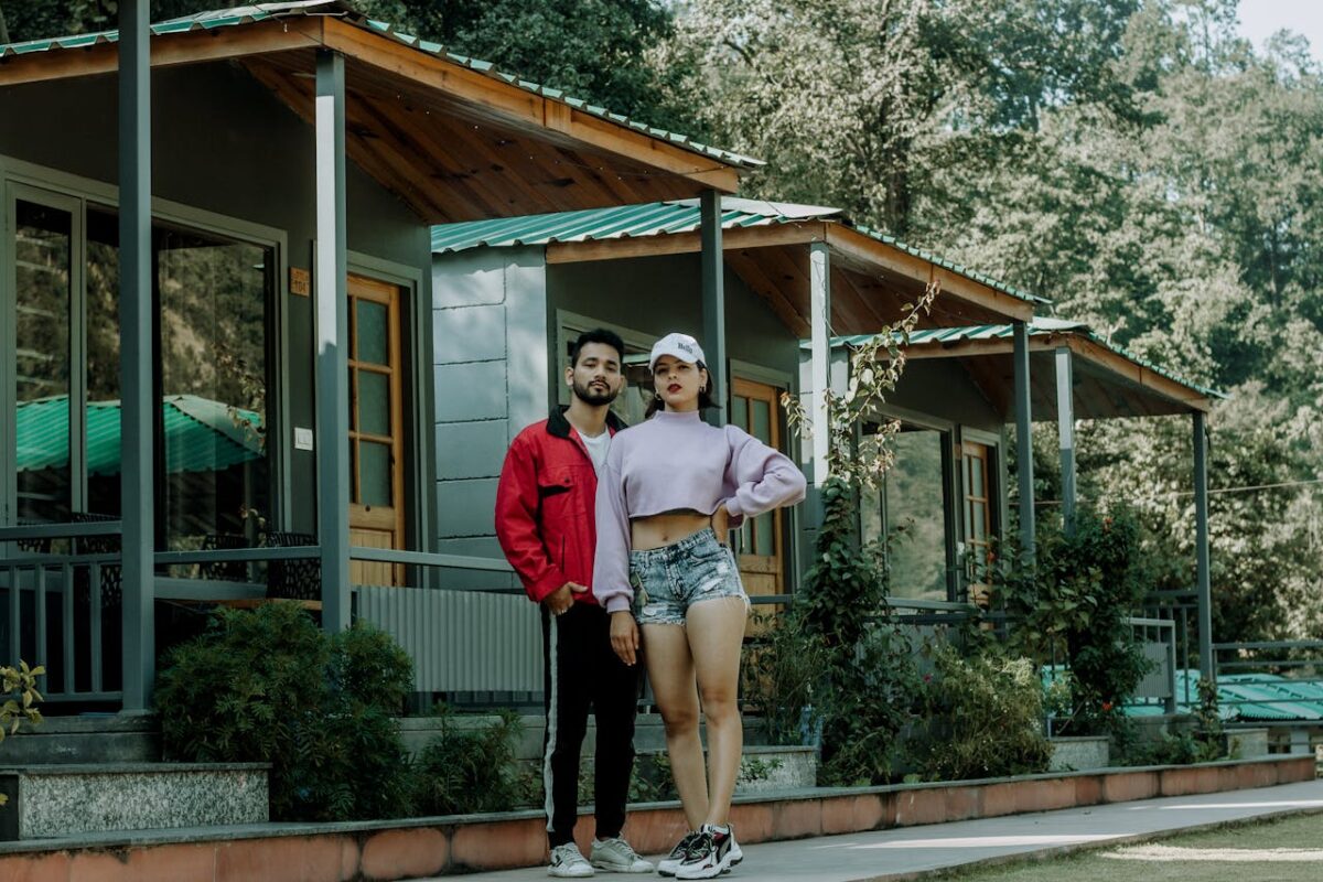 A stylish young couple poses outside a modern cabin surrounded by greenery.
