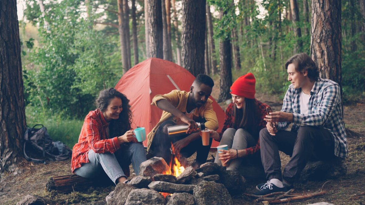 Group of friends gathered around a campfire at a forest campsite, enjoying drinks and conversation.